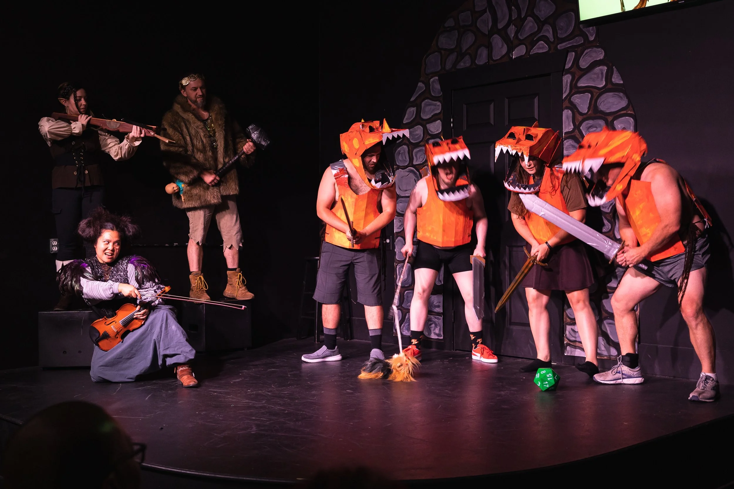 Theatrical performance featuring actors in costumes, including some dressed as dragons and others with medieval weapons, with a violinist and a violin on stage, set against a stone castle backdrop.