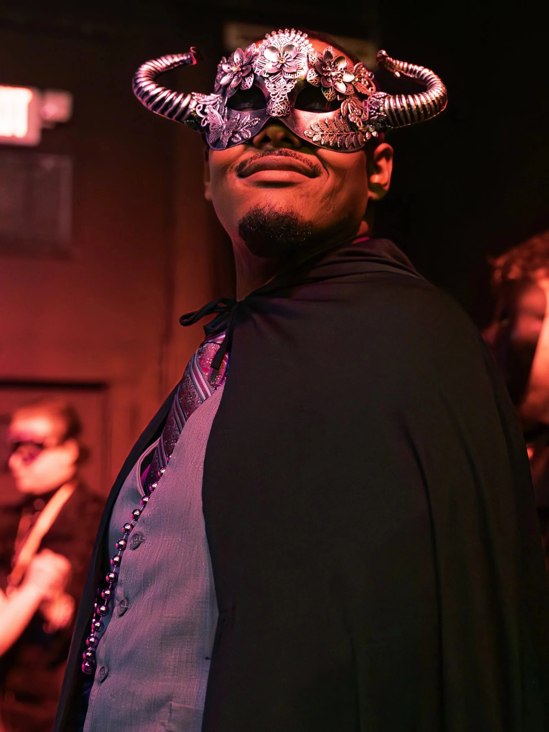 A man wearing an elaborate horned mask decorated with flowers and butterflies, smiling with their head turned to the side in a dimly lit setting.
