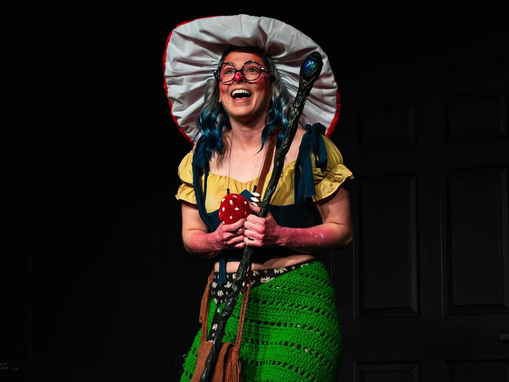 A woman dressed as a fairy tale character wearing a large white and red hat, glasses, a yellow top with blue accents, a green crochet skirt, and holding a magic staff and a mushroom toy, standing in front of a dark background, smiling and appearing joyful.