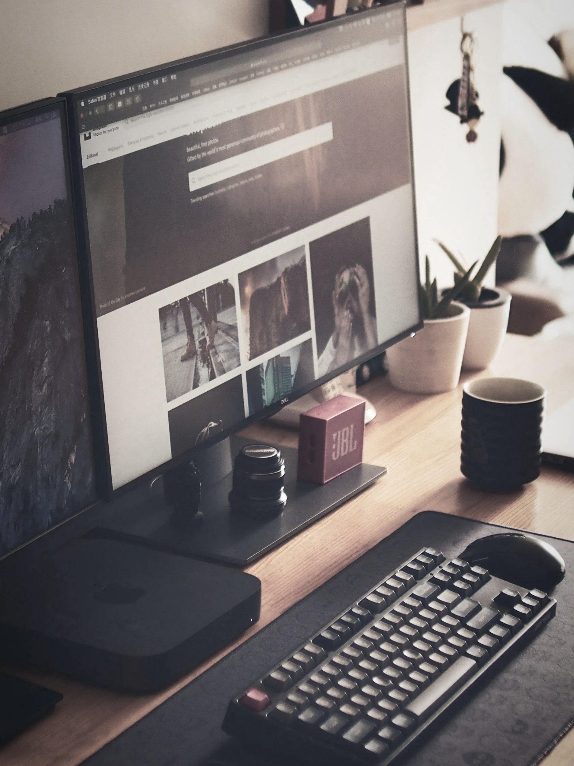 A computer desk with two monitors, one displaying a website and the other partially visible. There is a keyboard, mouse, and a laptop, along with various small objects like a JBL speaker, a small pink speaker, and several potted plants in the background. The setup is minimalist with a wooden surface.
