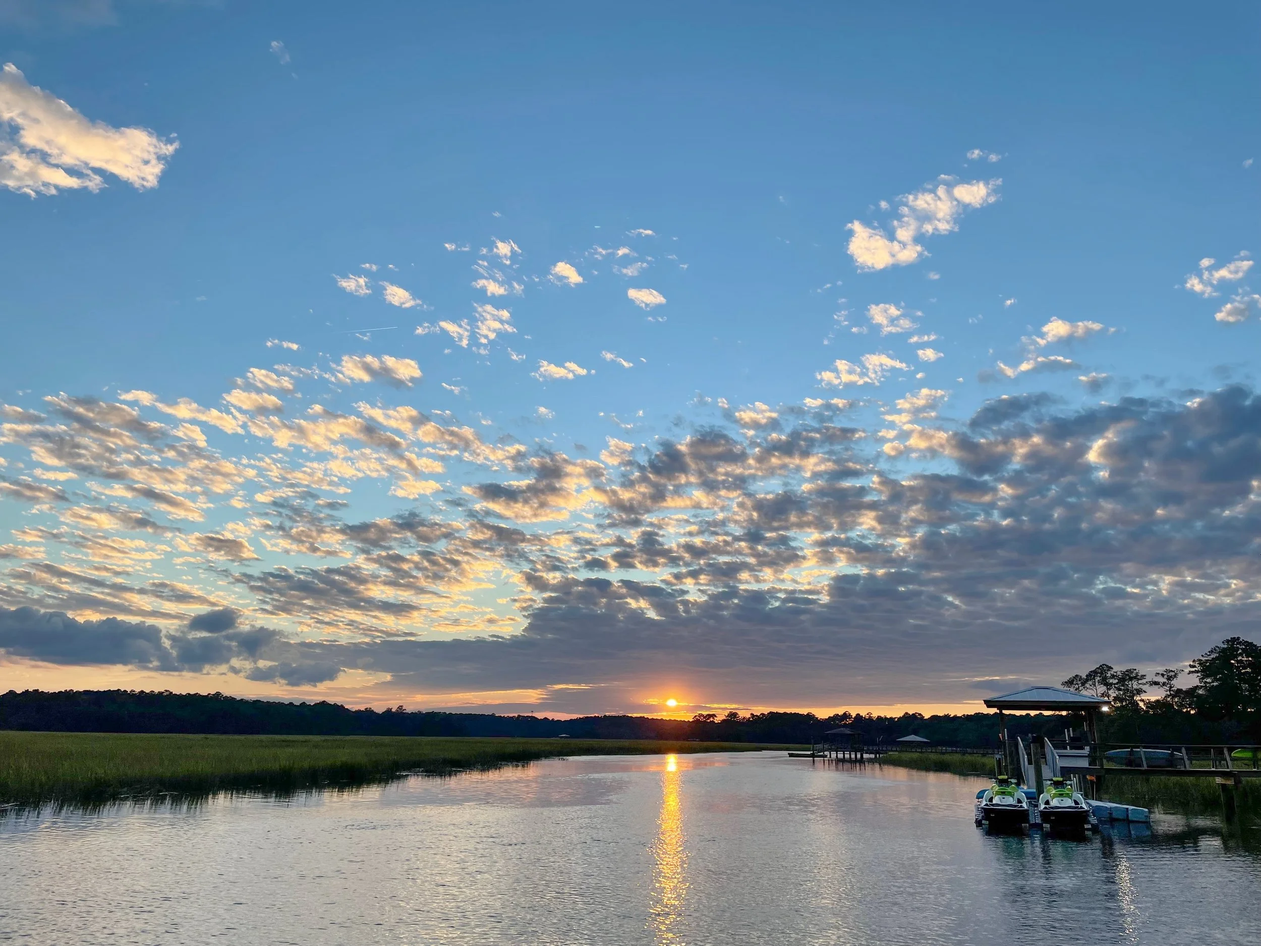 Sunset over a river with partly cloudy sky and a dock with small boats on the right side of the water.