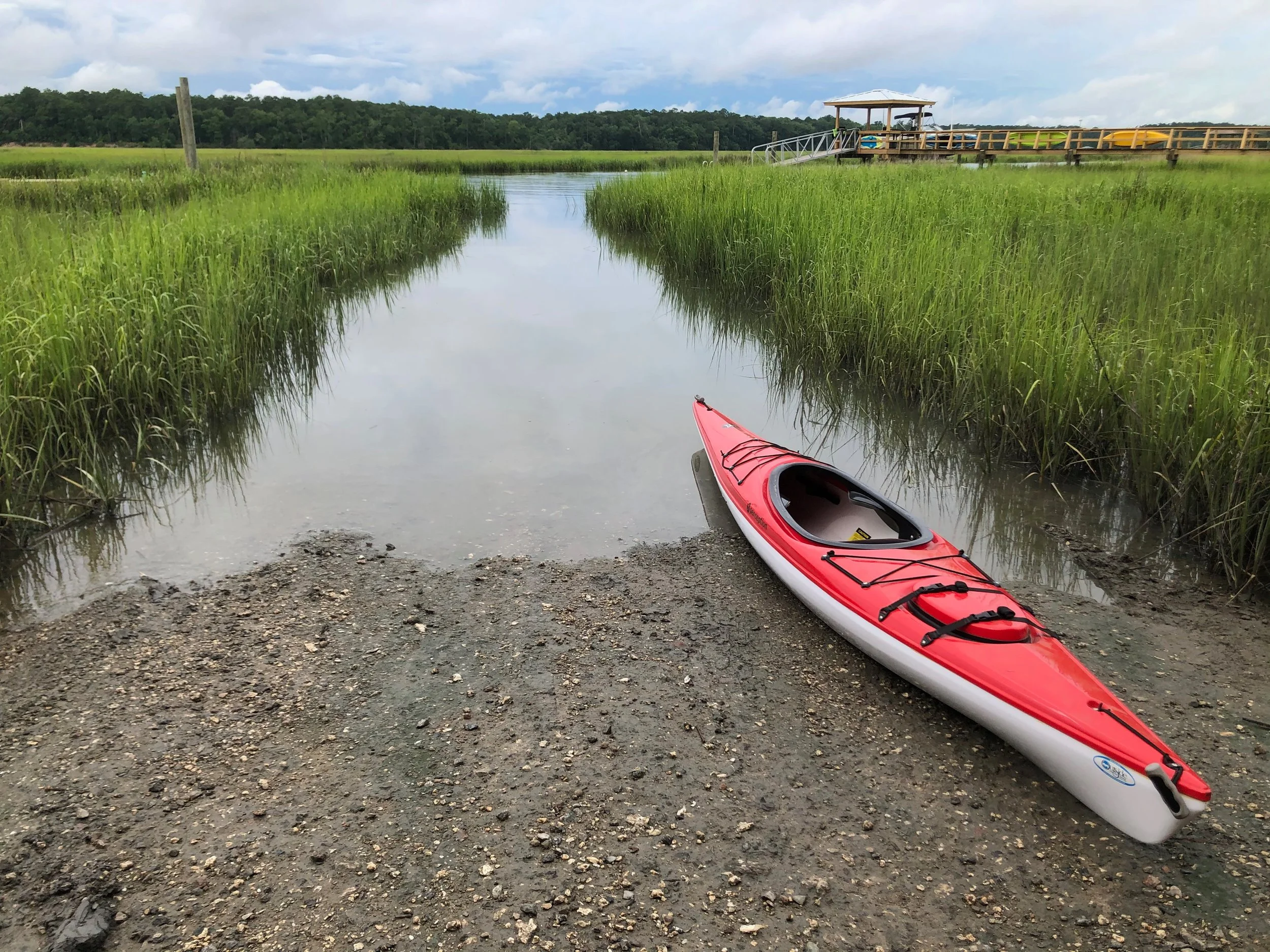 A red and white kayak resting on muddy ground at the edge of a calm marsh, with tall green grass and a wooden deck with yellow paddle boats in the background.