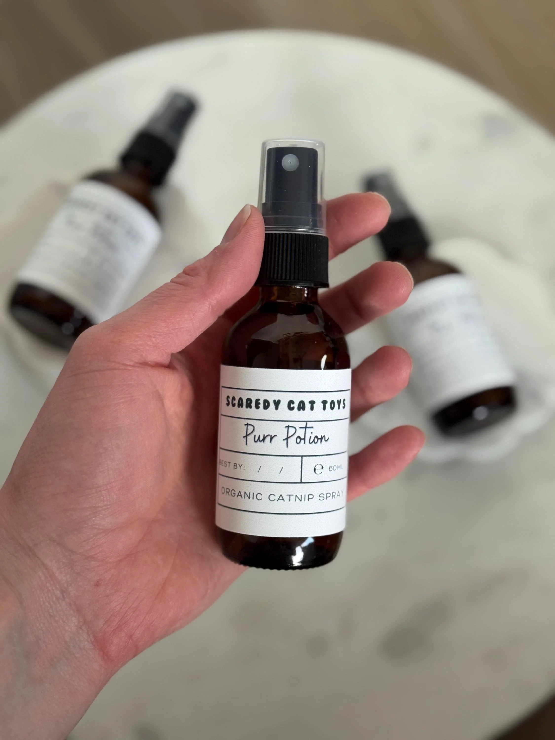 A person holding a small amber spray bottle labeled 'Scaredy Cat Toys Purr Potion Organic Catnip Spray' with two other bottles in the background on a white surface.