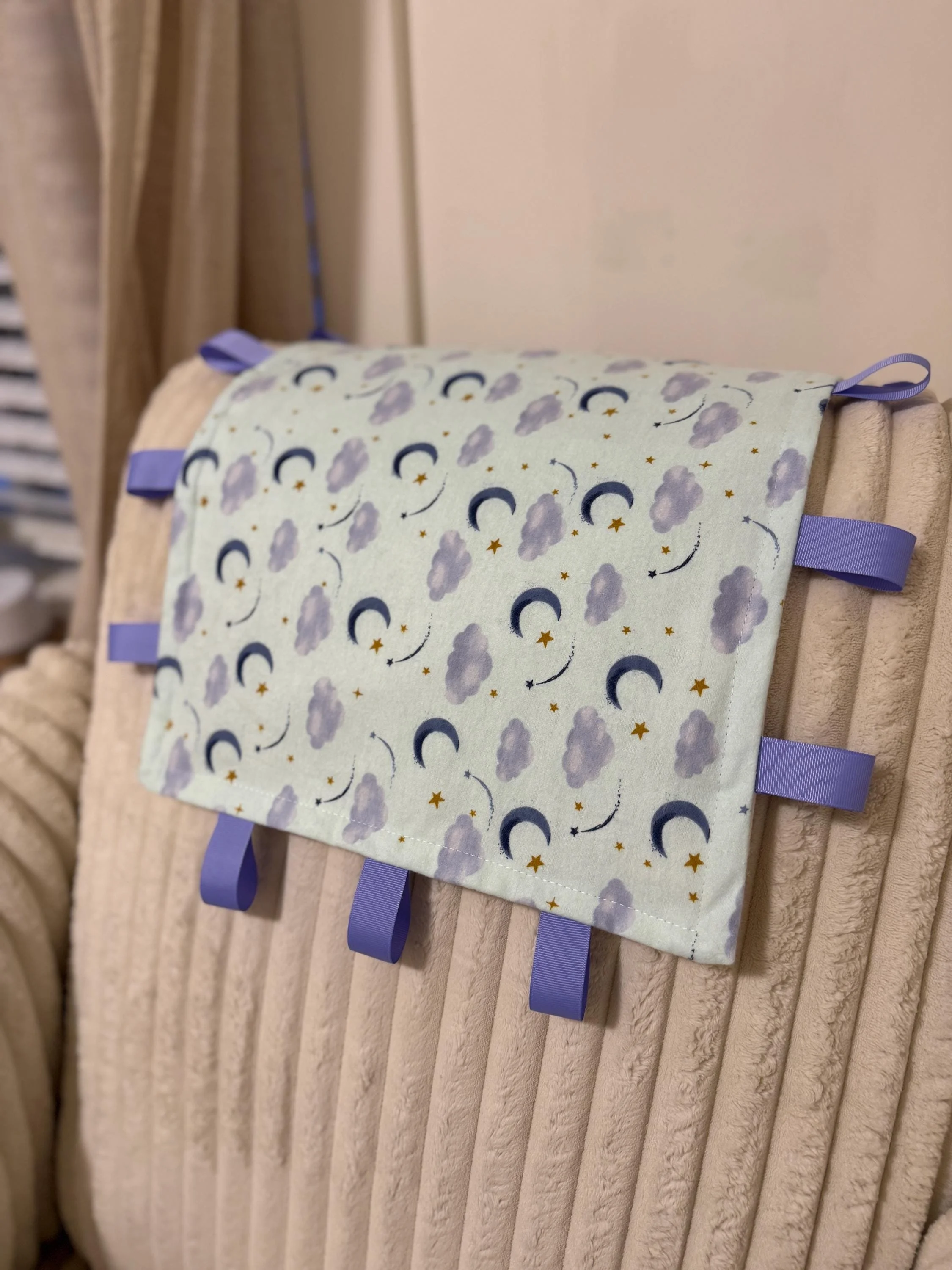 A crinkle mat with a print of clouds, moons, and stars. The mat is bordered with blue ribbon loops. draped over a beige, textured cushion or bed.