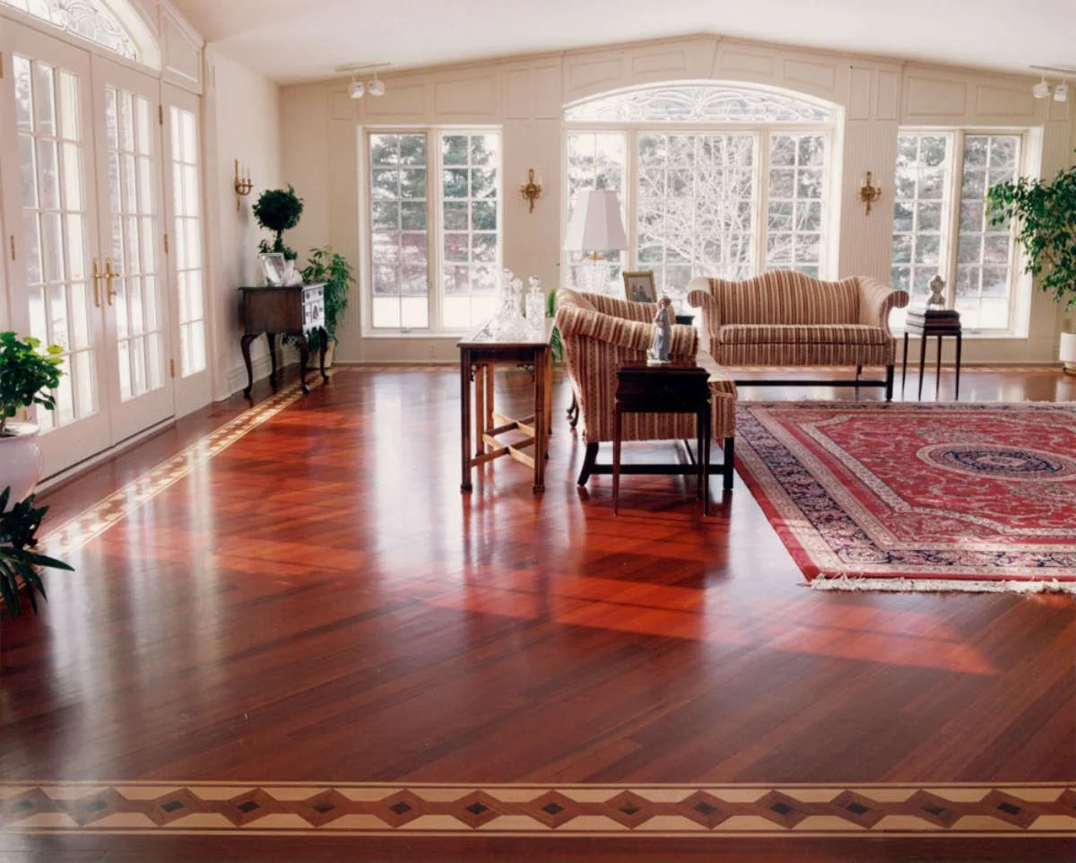 A spacious living room with hardwood floors, large windows, a sofa, armchairs, a red patterned rug, and plants.