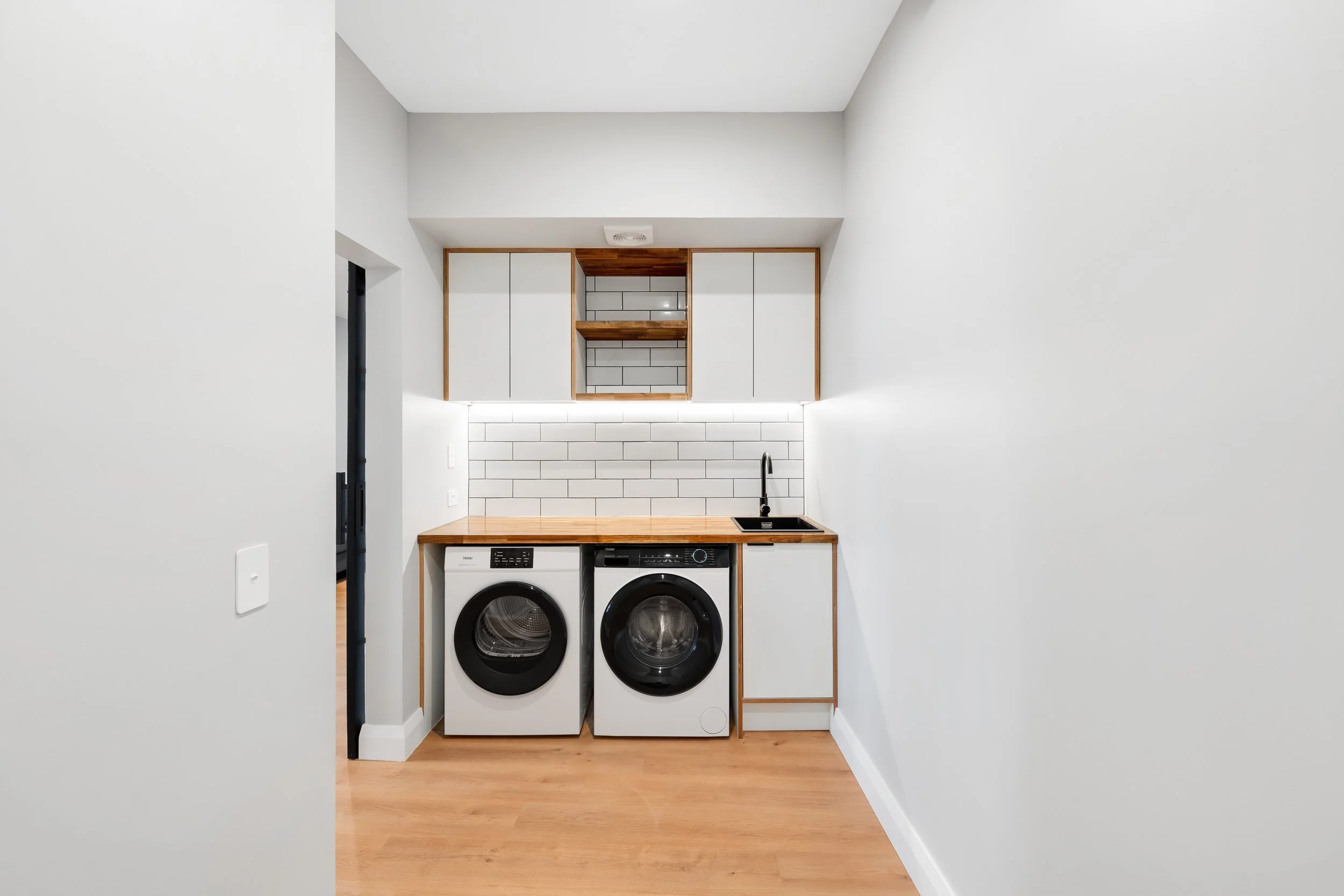 Small laundry room with white walls, wooden countertop, washing machine and dryer, white subway tile backsplash, black sink, and upper cabinets with open shelving.