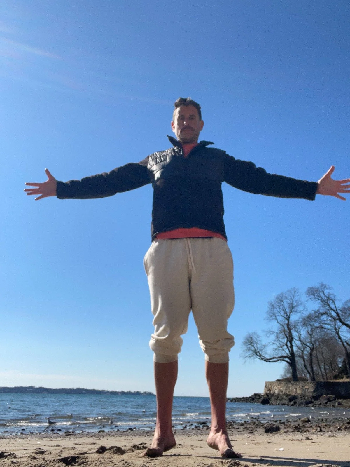 A man standing on a beach with arms outstretched, wearing a black jacket and beige shorts, with trees and water in the background under a clear blue sky.