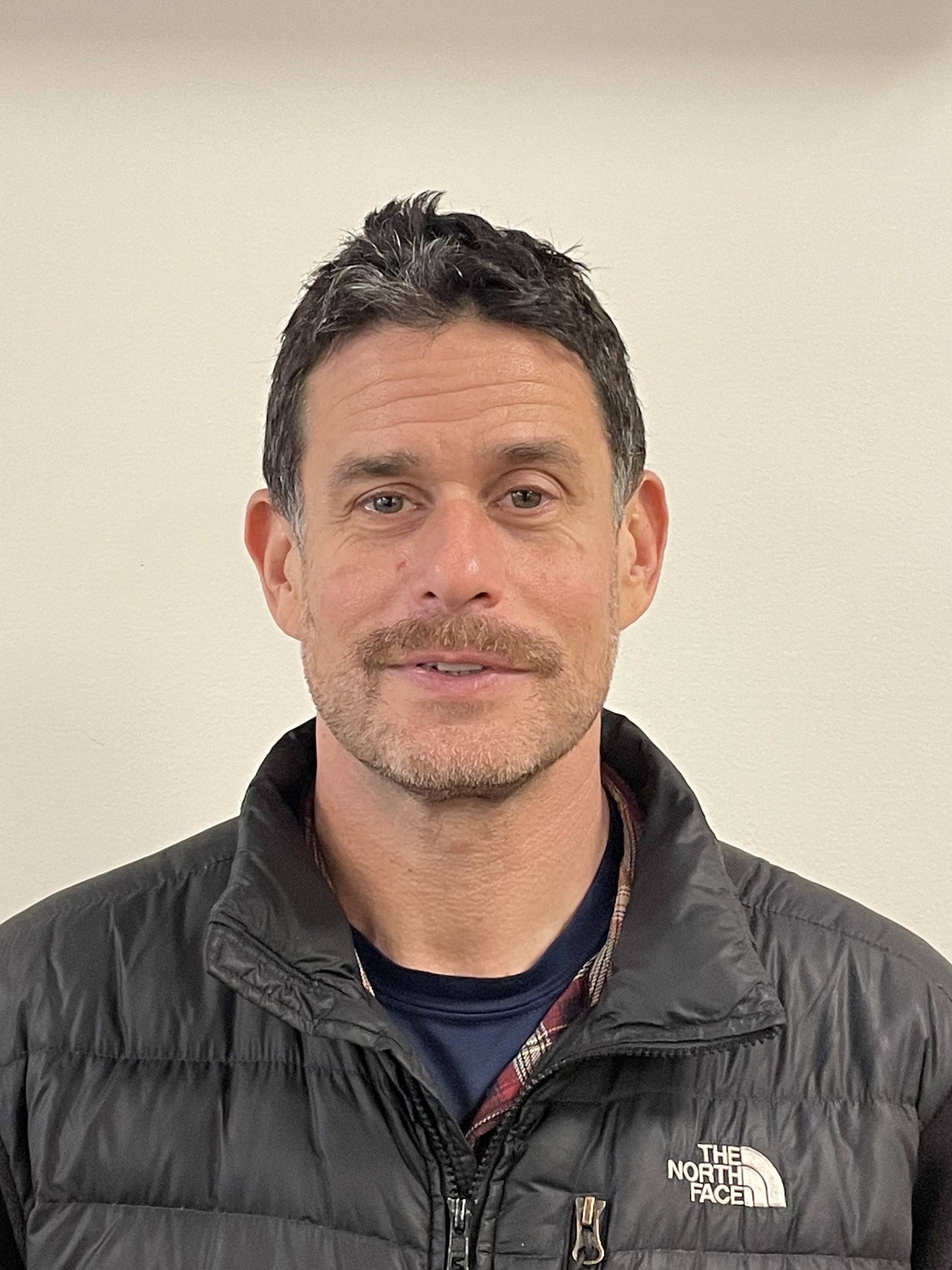 A close-up photo of a middle-aged man with dark, curly hair and a light beard, wearing a black North Face puffer jacket, standing against a plain cream wall background.