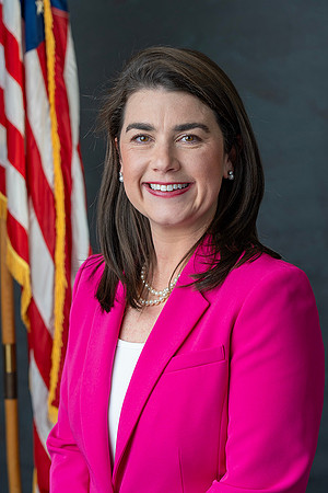 A woman with dark hair wearing a bright pink blazer, white top, pearl necklace, and pearl earrings, standing in front of an American flag and a dark background.