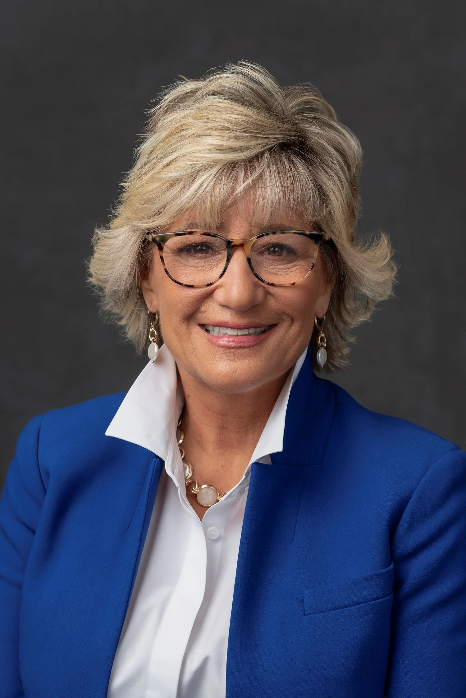 Portrait of a smiling middle-aged woman with blonde hair, wearing glasses, a blue blazer, and a white shirt, against a dark background.