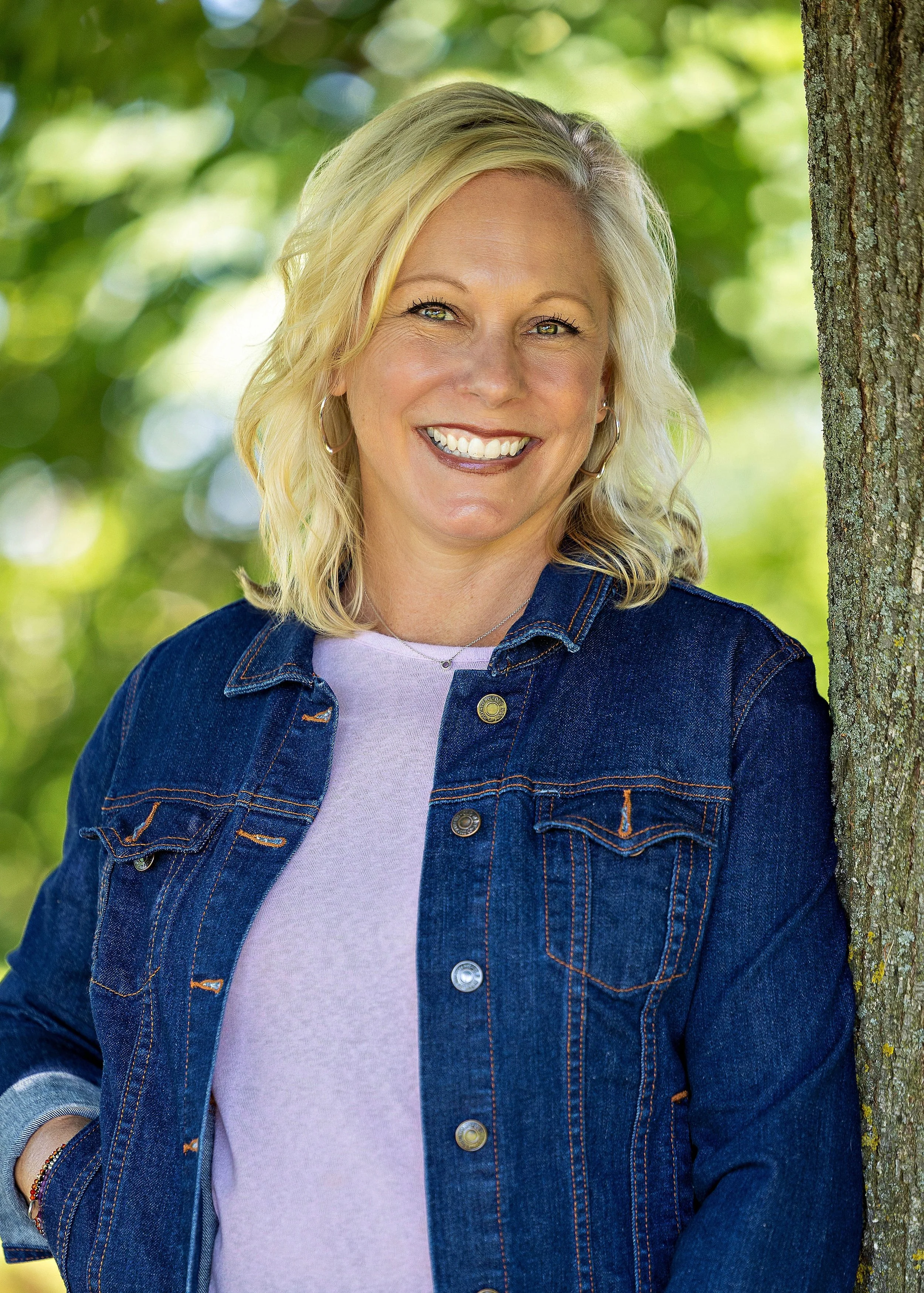 A smiling woman with blonde hair, wearing hoop earrings, a light purple shirt, and a dark denim jacket, standing outdoors in front of a tree with a blurred green background.
