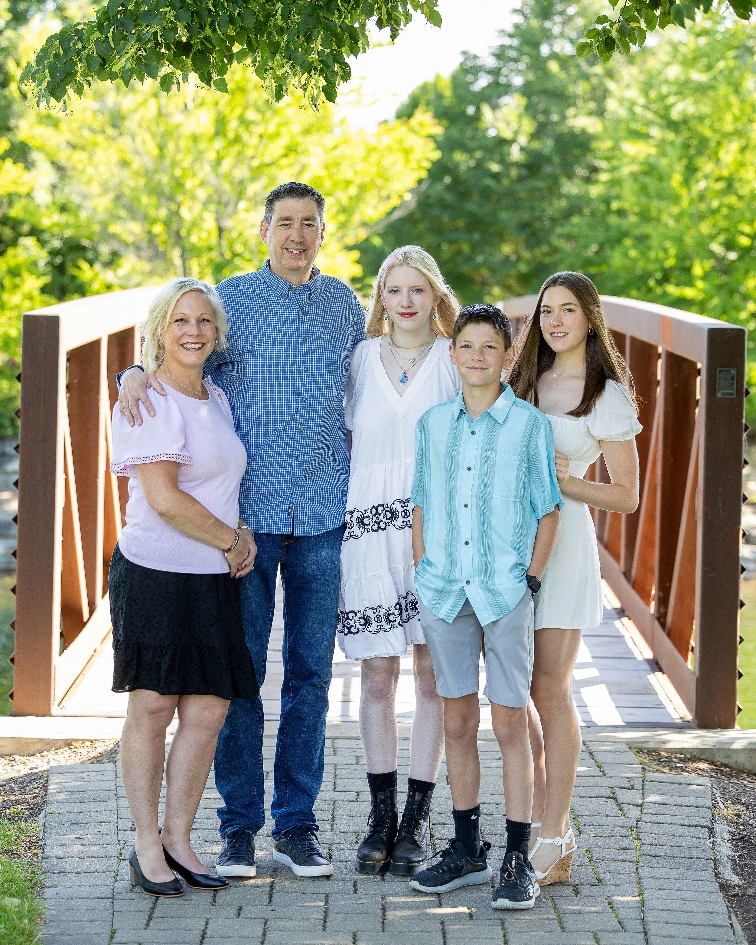 A family of five standing on a walkway on a bridge with green trees in the background. They are smiling and casually dressed, with some wearing dresses, shorts, or jeans.