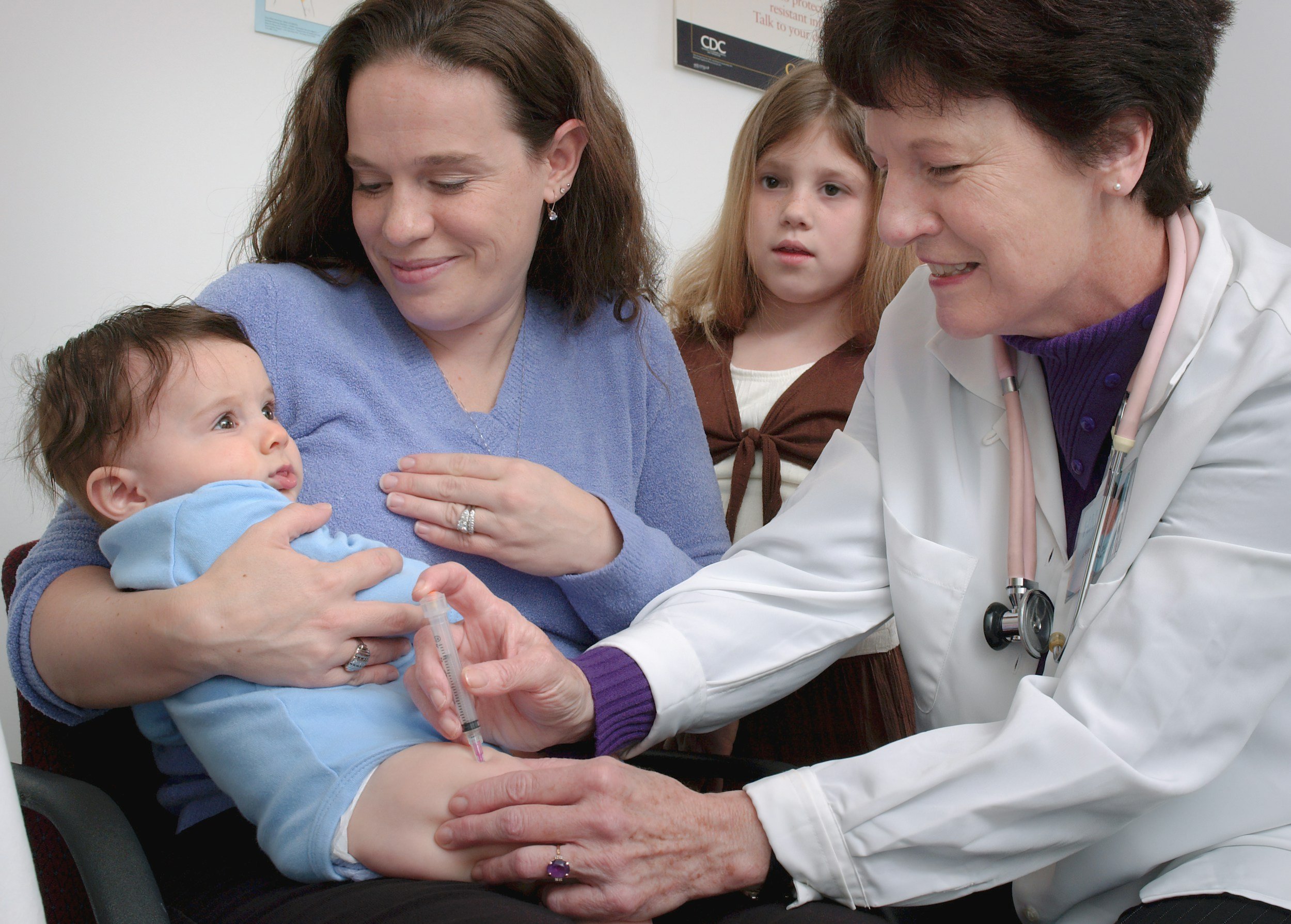 A doctor administering a vaccine to an infant girl while a mother, two young girls, and a healthcare worker are present.