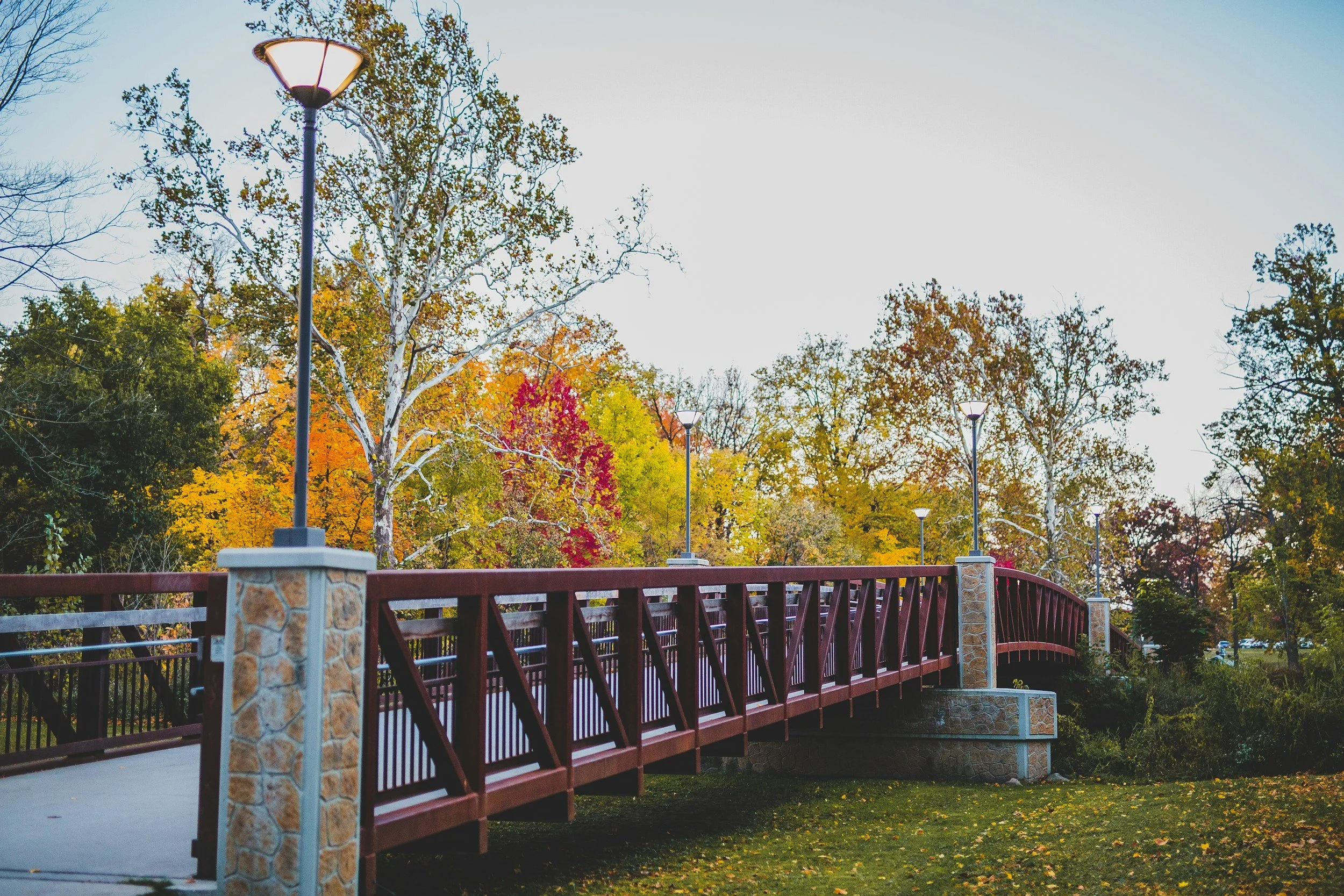 A red pedestrian bridge with stone pillars and lamp posts spans over a grassy area in a park with fall foliage trees in the background.