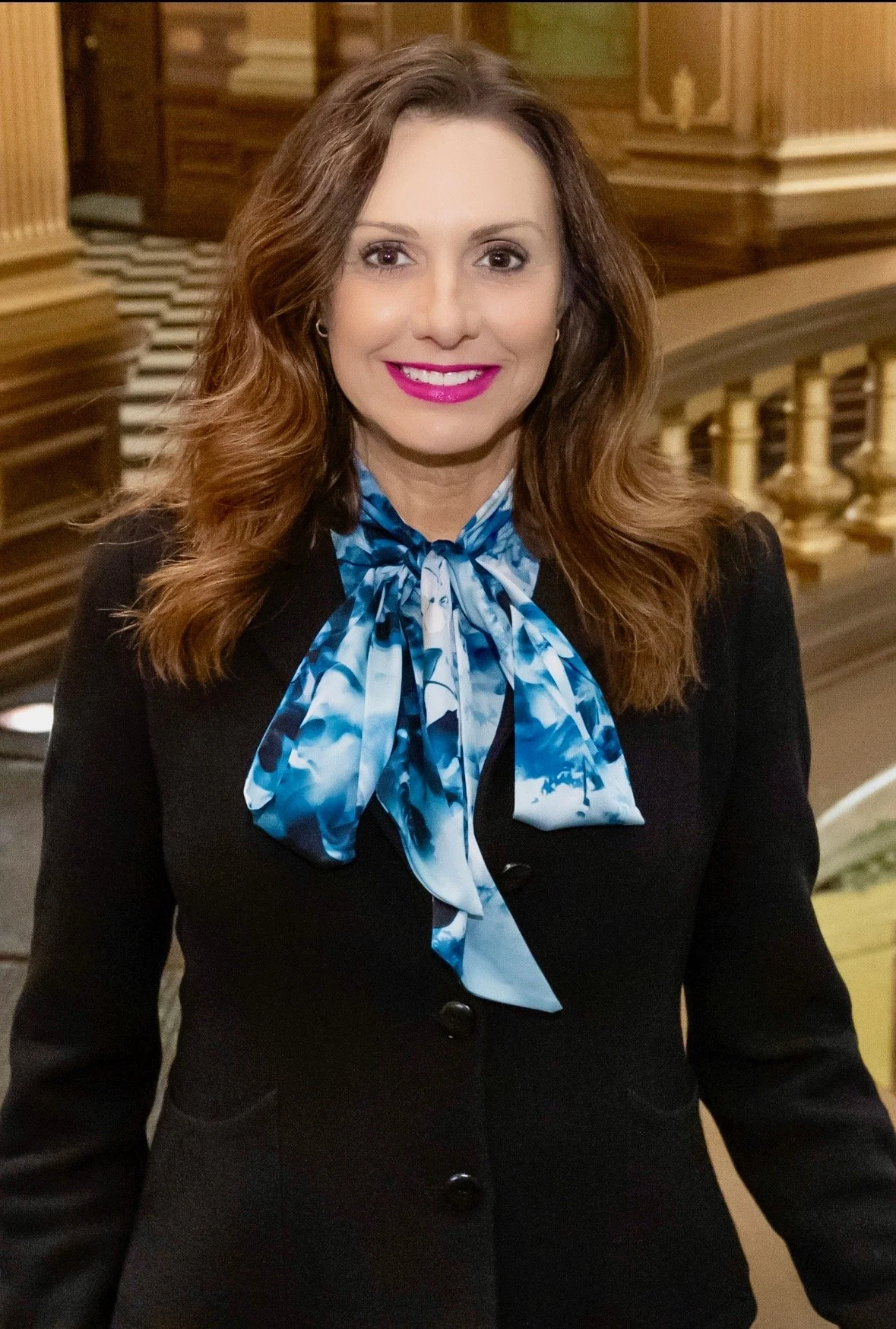 A woman with long brown wavy hair, pink lipstick, wearing a black blazer and a blue patterned silk scarf tied in a bow around her neck, standing indoors with warm wood-paneled decor.