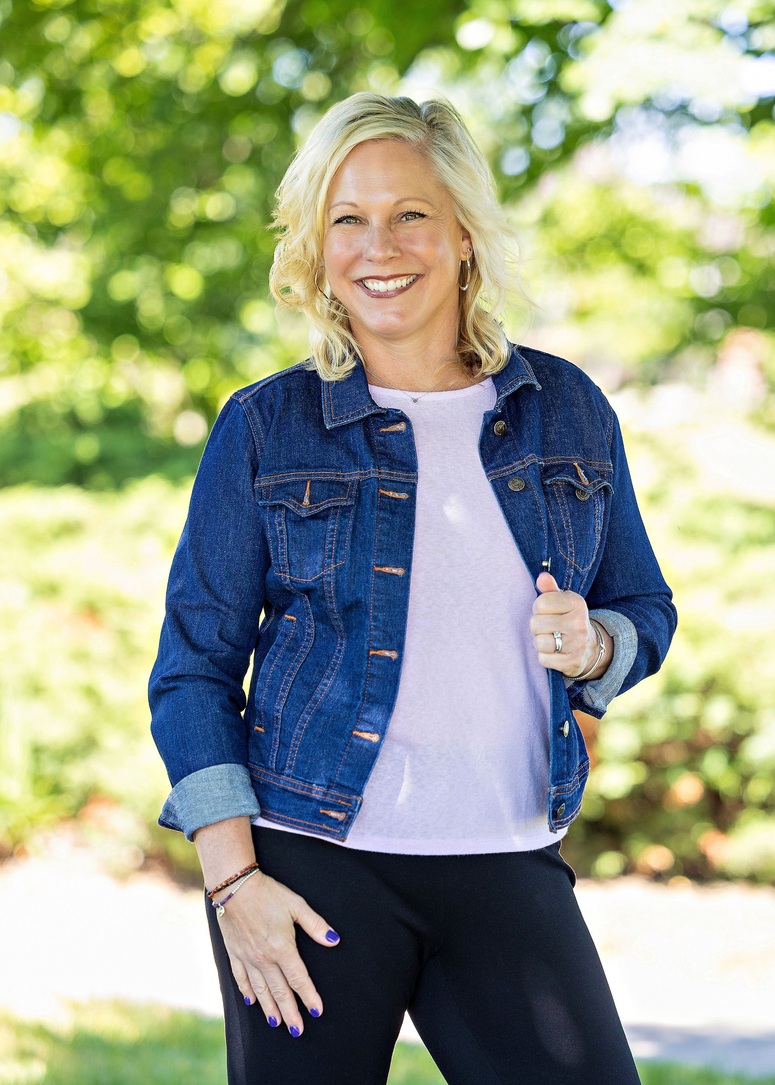 A smiling woman with blonde hair outdoors, wearing a denim jacket, a light purple shirt, and black pants, with a background of green trees and sunlight.