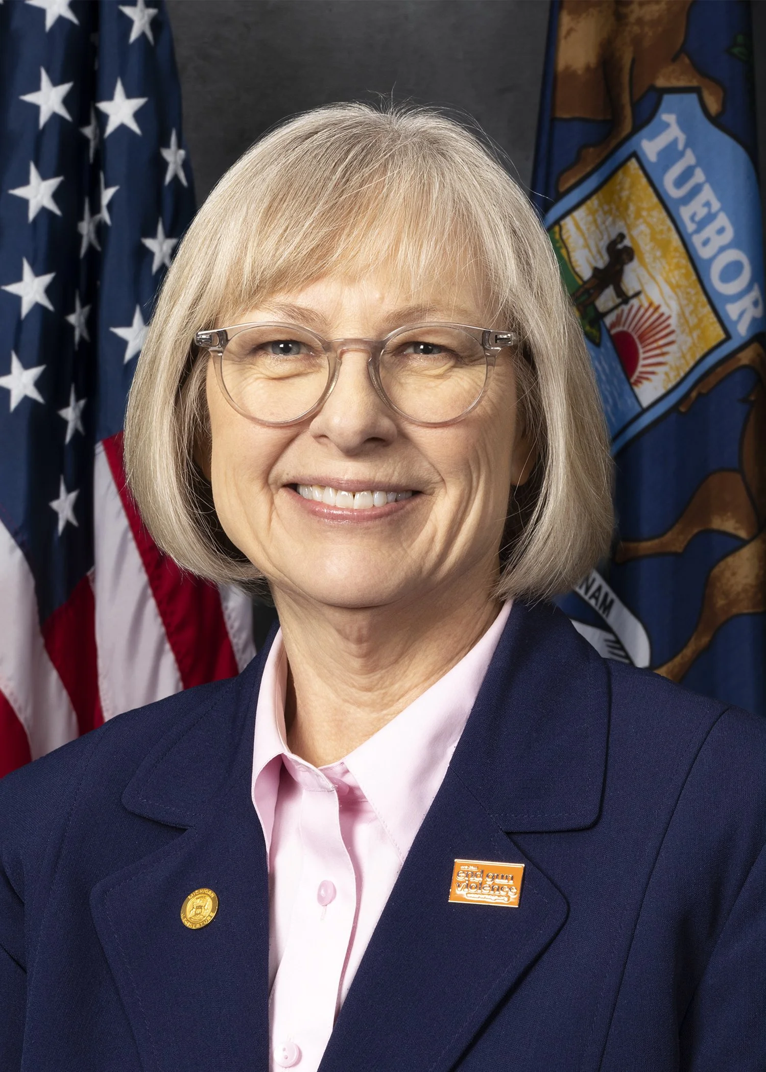 A smiling woman with blonde hair, wearing glasses, a navy blazer, and a pink shirt. Behind her are the American flag and a state flag of Nebraska.