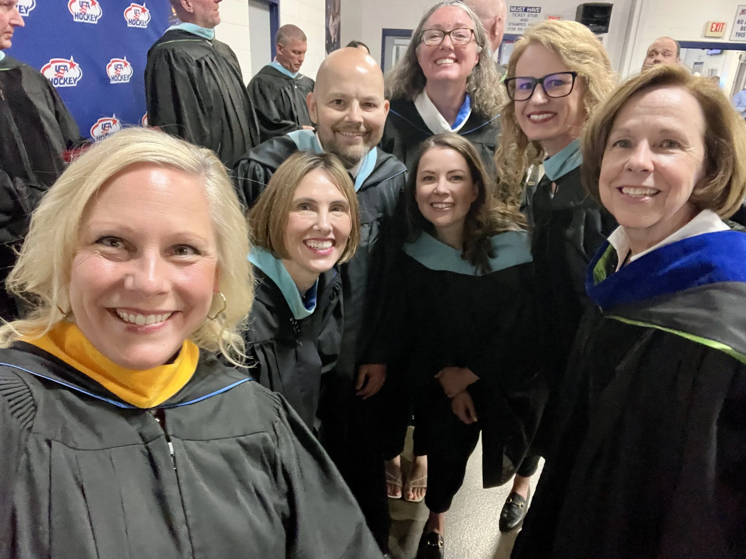 Group of people in graduation gowns and caps celebrating indoors, with a backdrop displaying 'USA Hockey' logos, indicating a graduation or award ceremony related to Northville Public Schools.