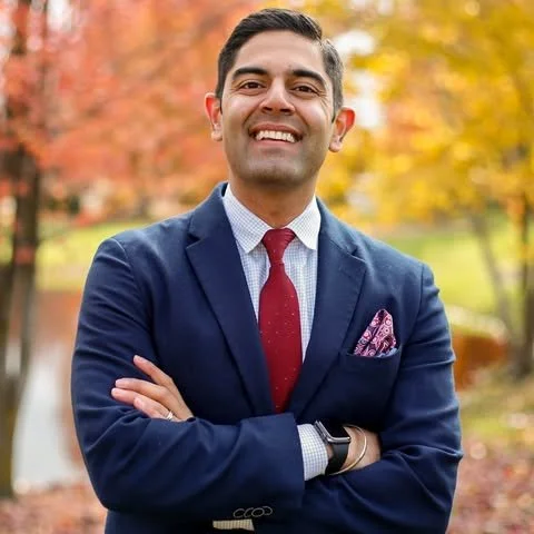 Smiling man in a navy suit, white shirt, and red tie standing outdoors with autumn trees in the background.