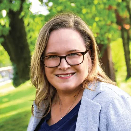 A woman with shoulder-length brown hair, wearing glasses, a light gray blazer, and a navy shirt, smiling outdoors with green trees and grass in the background.