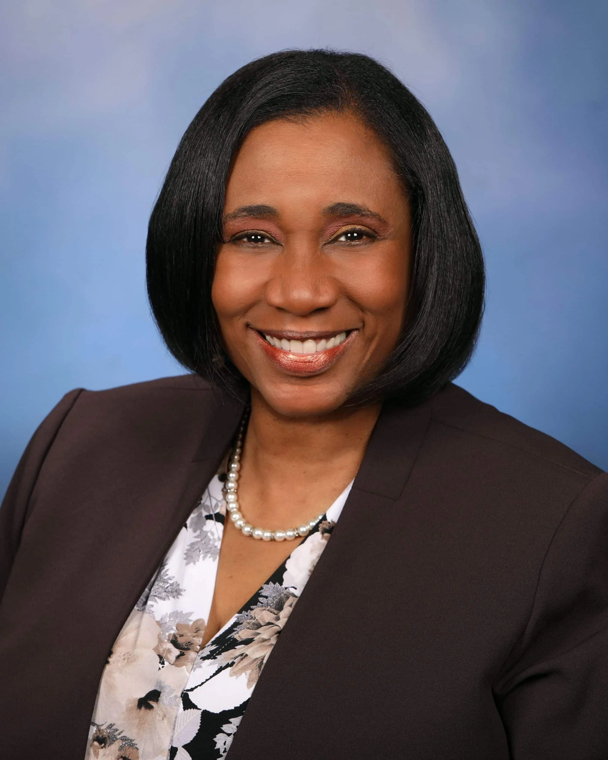 A professional woman with a bob hairstyle, wearing a black blazer, floral blouse, pearl necklace, and smiling against a blue background.