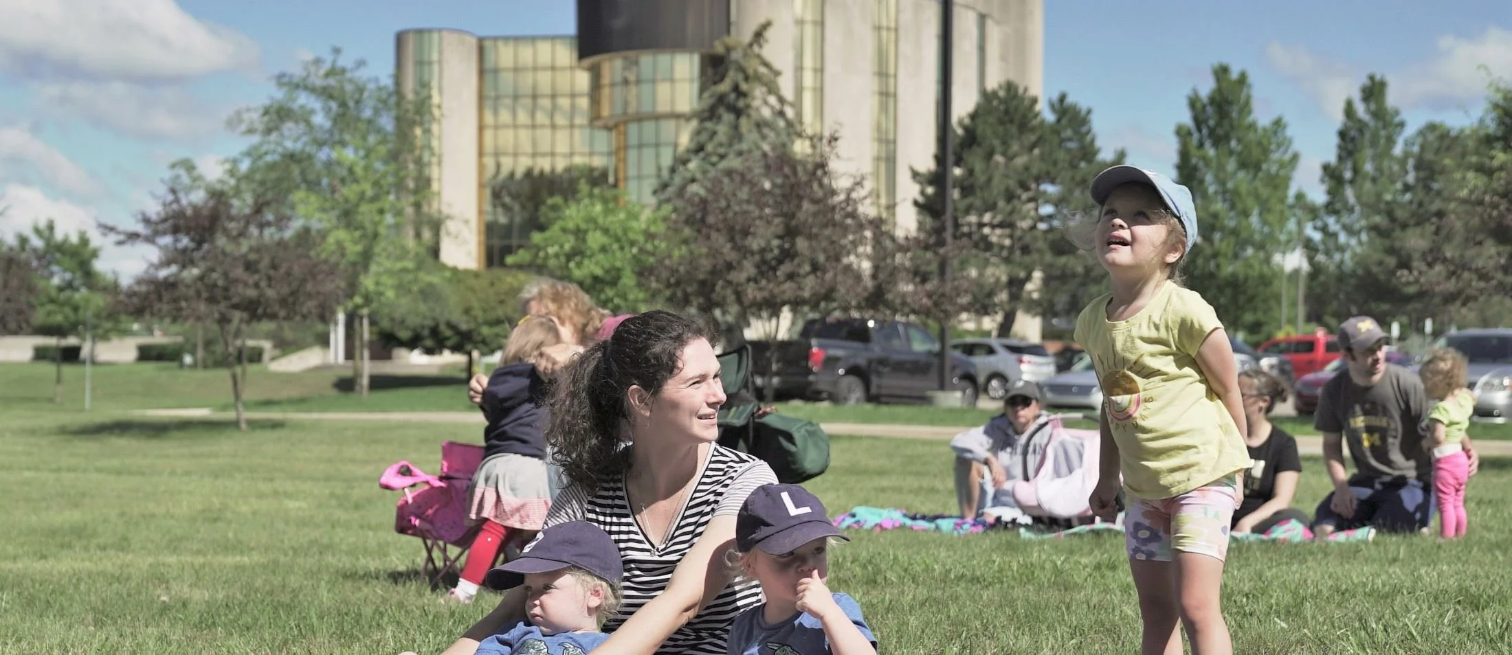 A group of people, including children and adults, enjoying a sunny day at a park in Livonia with trees, grass, and a tall building in the background. A woman is sitting on the grass with two children, one wearing a blue cap, and a yellow shirt