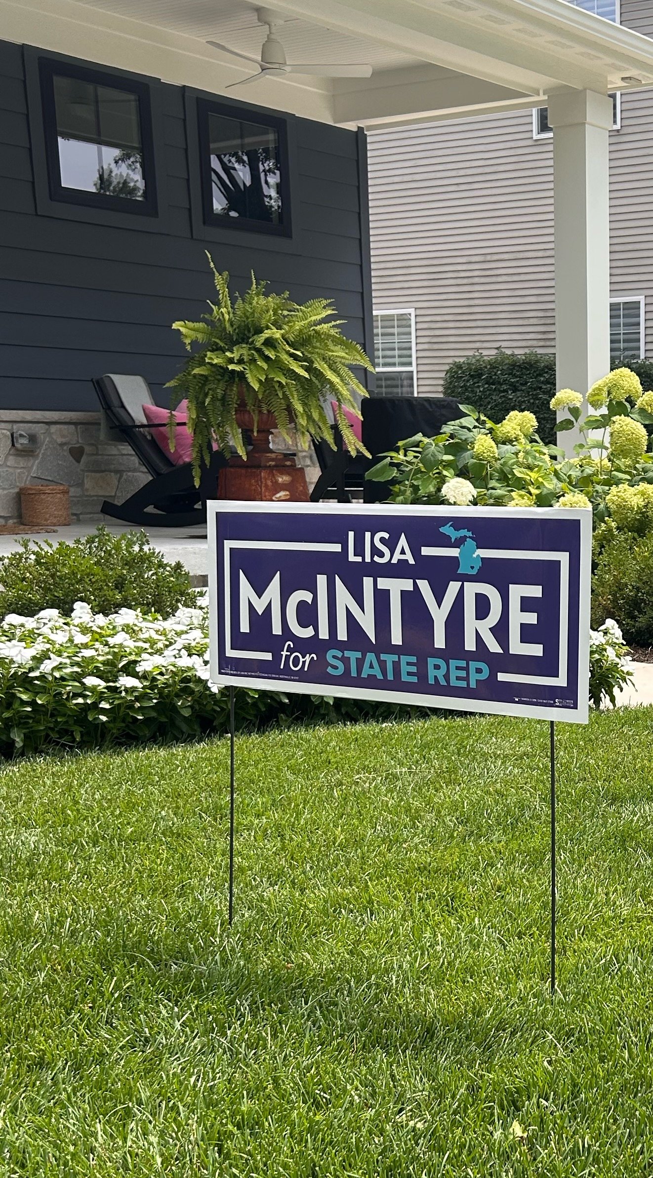 A campaign sign on a lawn supporting Lisa McIntyre for State Representative, with a house and garden in the background.
