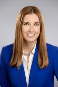 Professional woman with long straight red hair, wearing a blue blazer and white shirt, smiling against a neutral background.