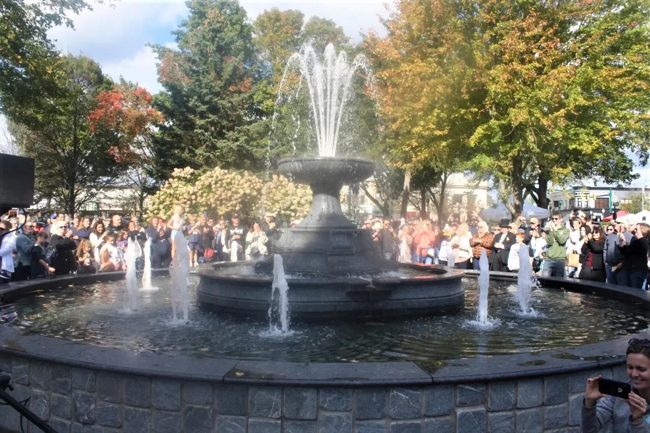 Crowd gathered around a fountain in a park in Plymouth MI with trees showing fall foliage.