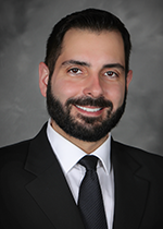 Headshot of a man with dark hair and beard, wearing a black suit and white shirt, smiling against a dark background.