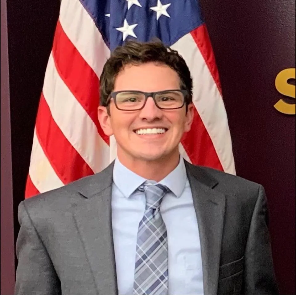 Smiling man in glasses and a gray suit standing in front of an American flag.
