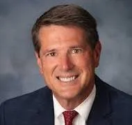 Professional headshot of a smiling man with brown hair, wearing a dark suit, white shirt, and red tie, against a blue-gray background.