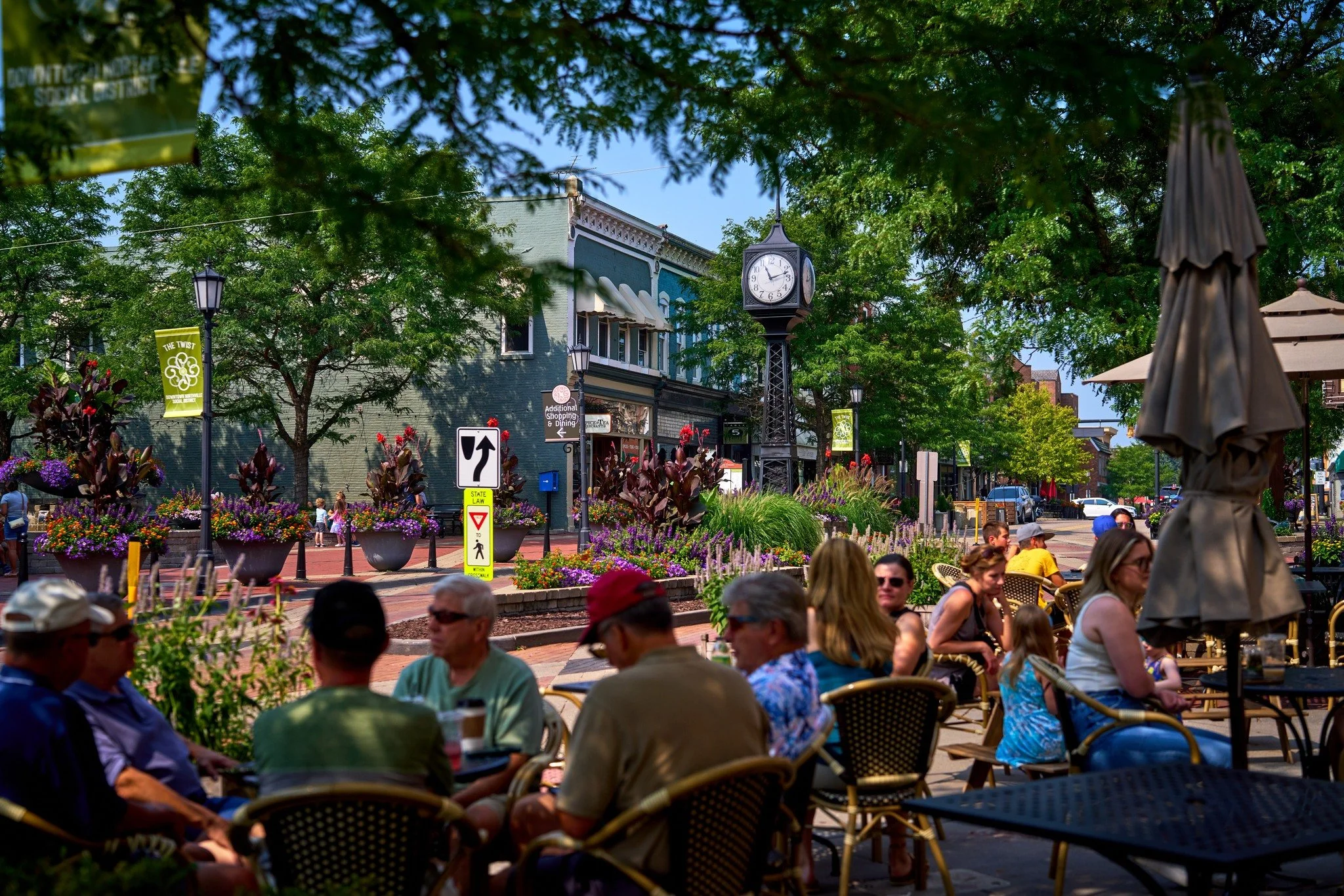 People dining outdoors in Northville, MI on a sunny day at a street cafe with colorful flowers, trees, and historic buildings in the background.