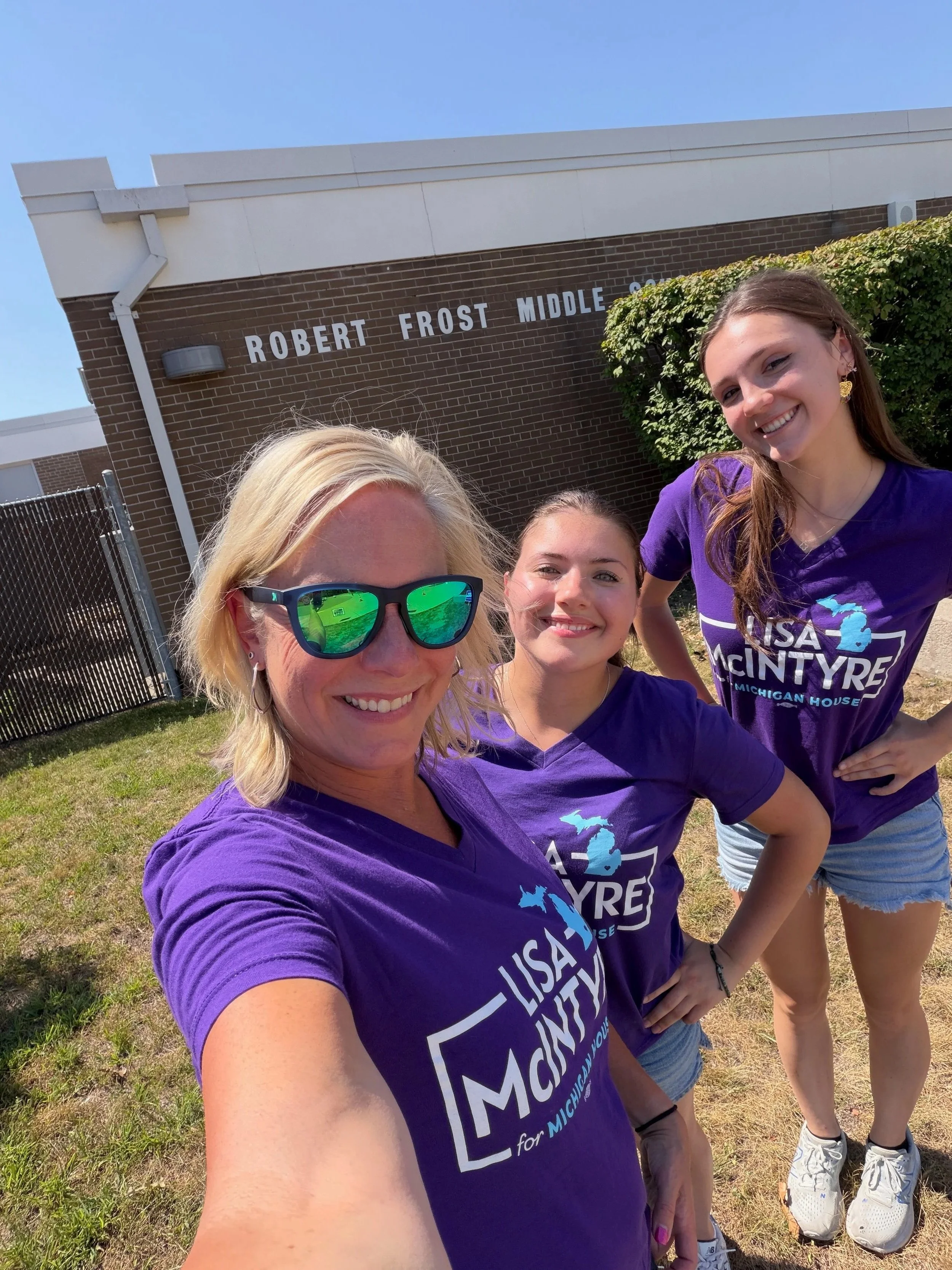 Four women taking a selfie outside chool. They are all wearing purple T-shirts with 'Lisa McIntyre for Michigan House' printed on them. The women are smiling, standing on grass, with a brick building and greenery in the background, under clear blue sky.