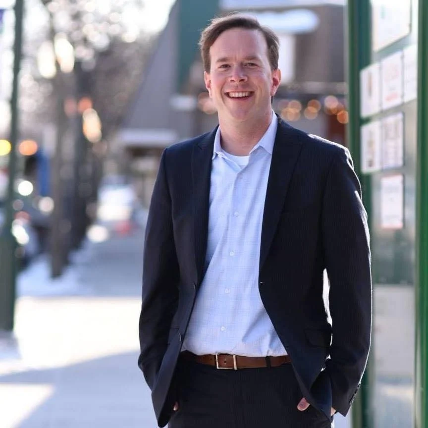 A young man wearing a dark suit jacket and light blue shirt smiling outdoors in a city setting.