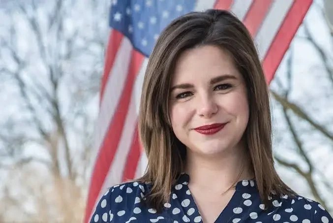 Young woman with brown hair and red lipstick standing outdoors in front of an American flag and trees.