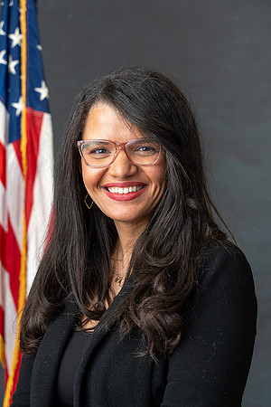 A woman with long dark hair and glasses smiling in front of an American flag.