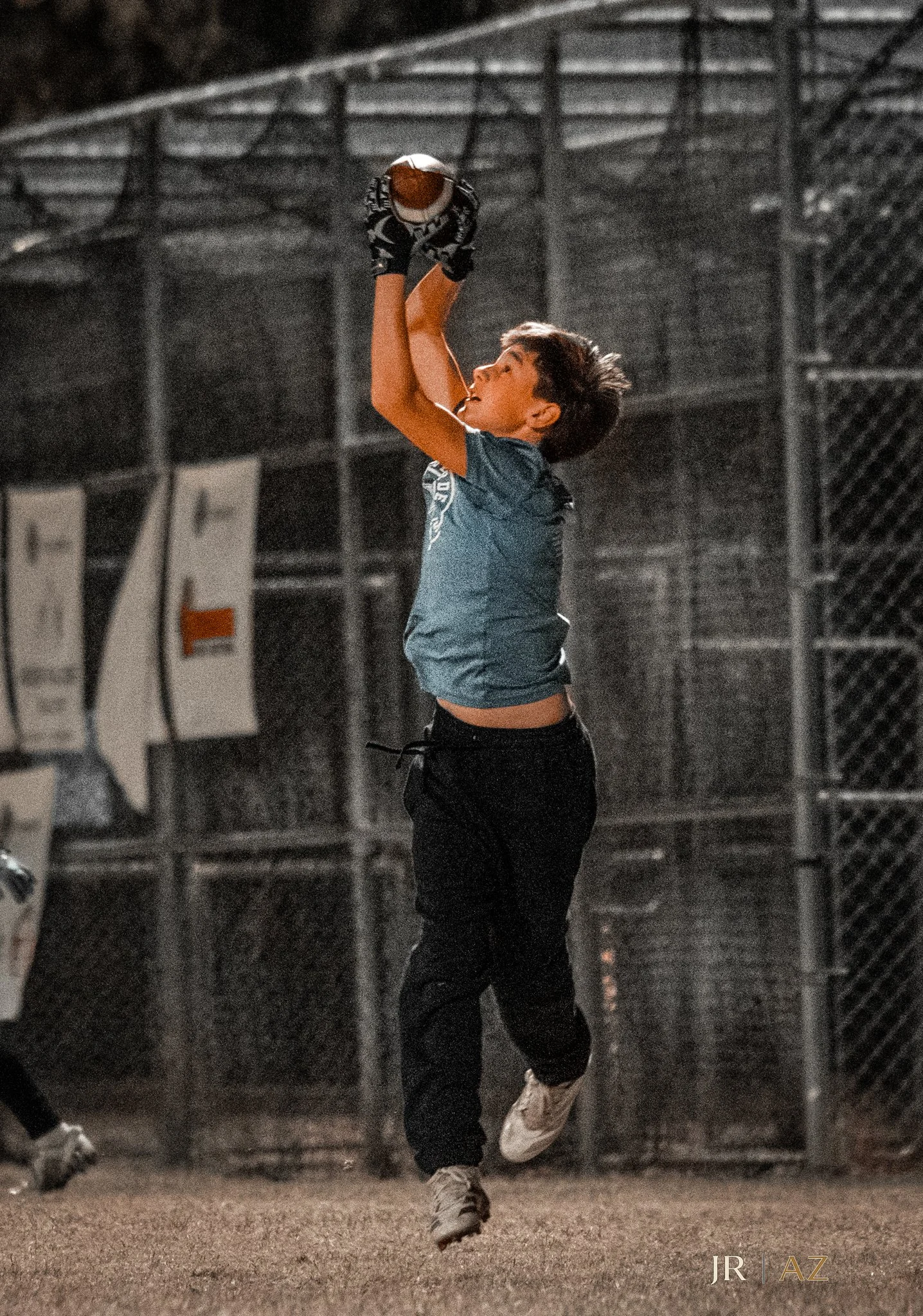 A young boy playing baseball at night, jumping to catch a ball with his glove in mid-air on a baseball field with a chain-link fence in the background.