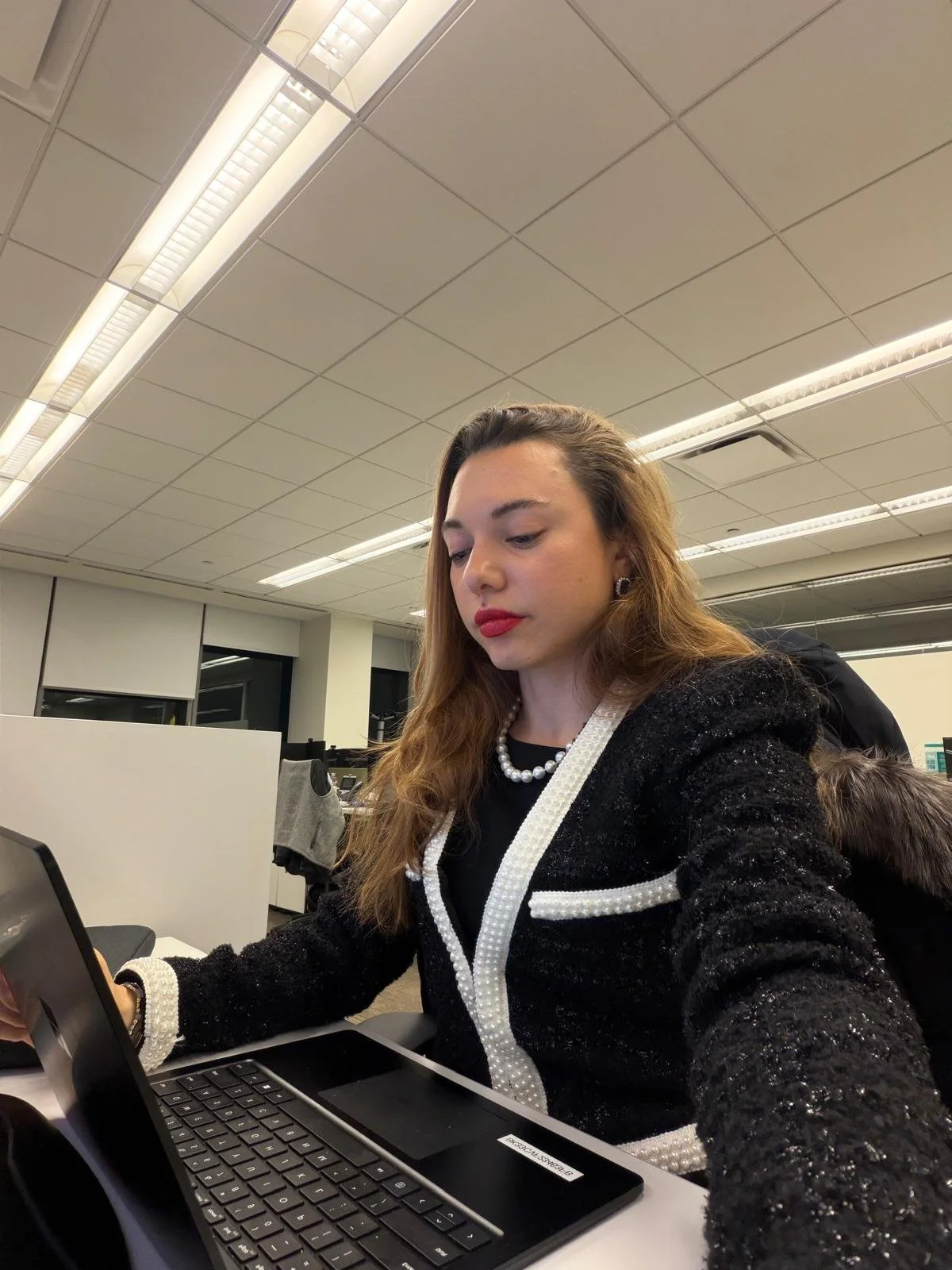 A woman sitting at a desk in an office, using a laptop, wearing a black and white outfit with pearl accents and red lipstick.