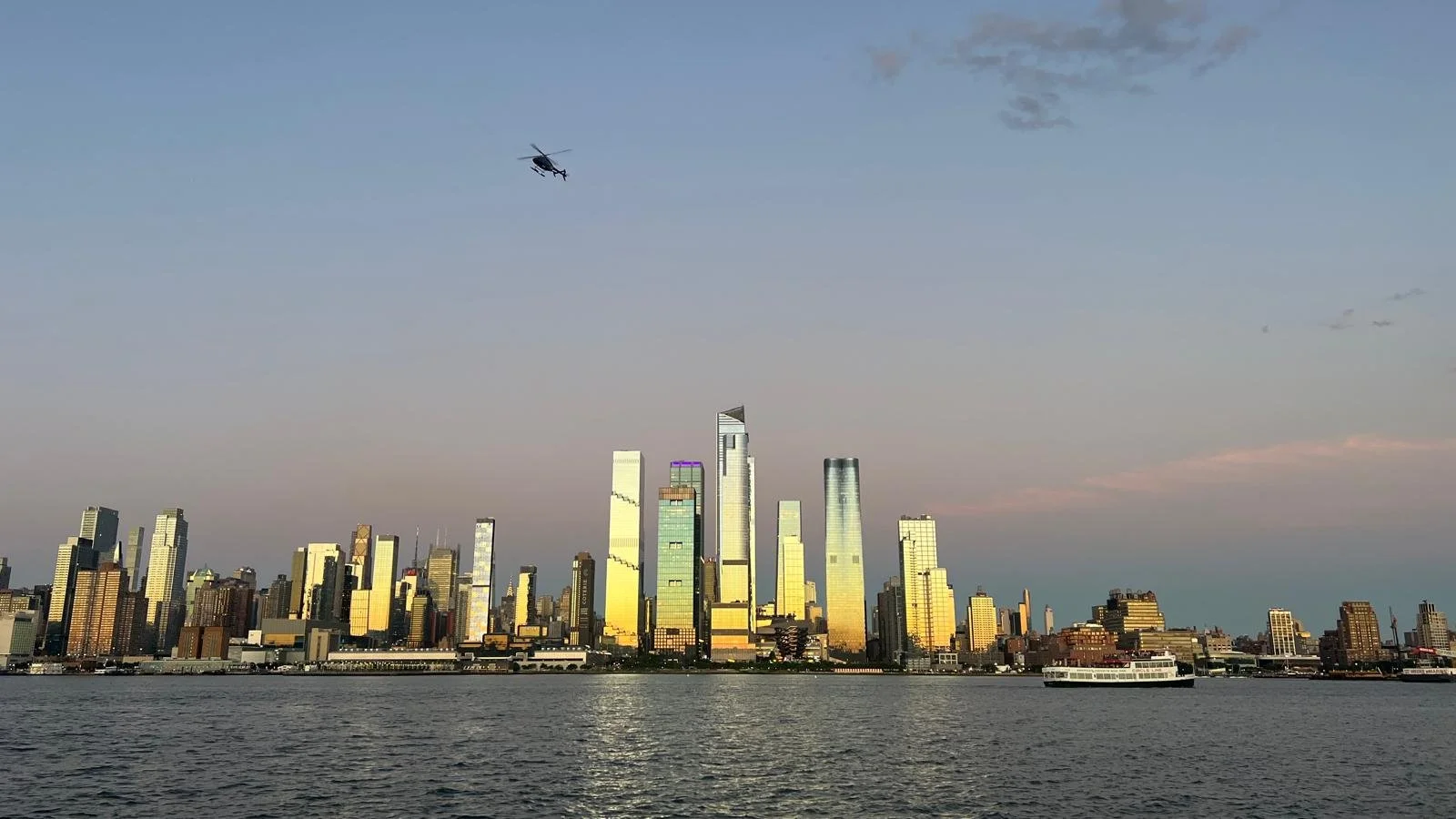 Skyline of New York City with skyscrapers, a boat on the river, and a helicopter flying overhead at sunset.
