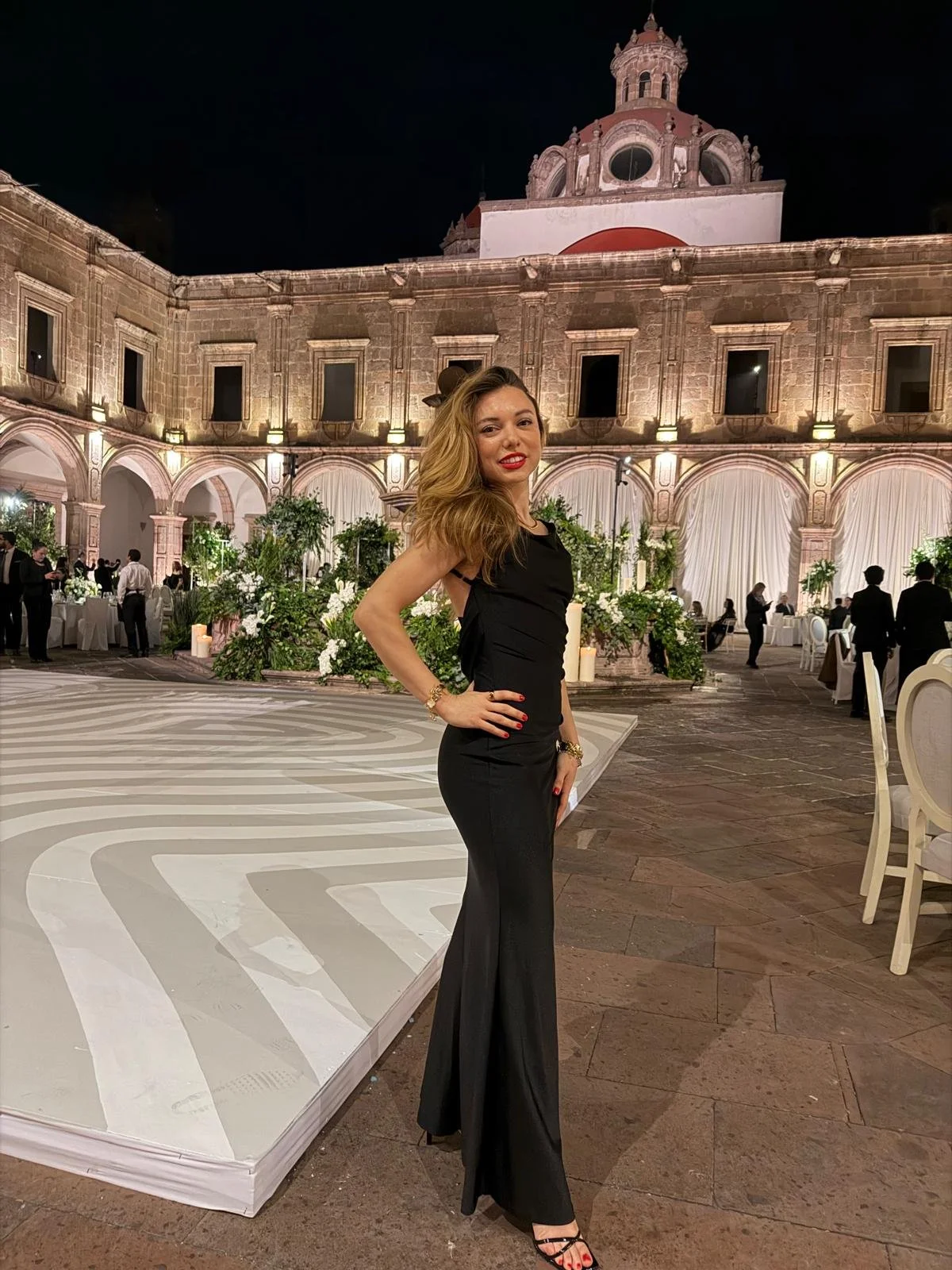 A woman in a black dress and high heels posing at an elegant outdoor event at night. The background features a historic building with illuminated arches and a domed tower, decorated with greenery and white flowers.