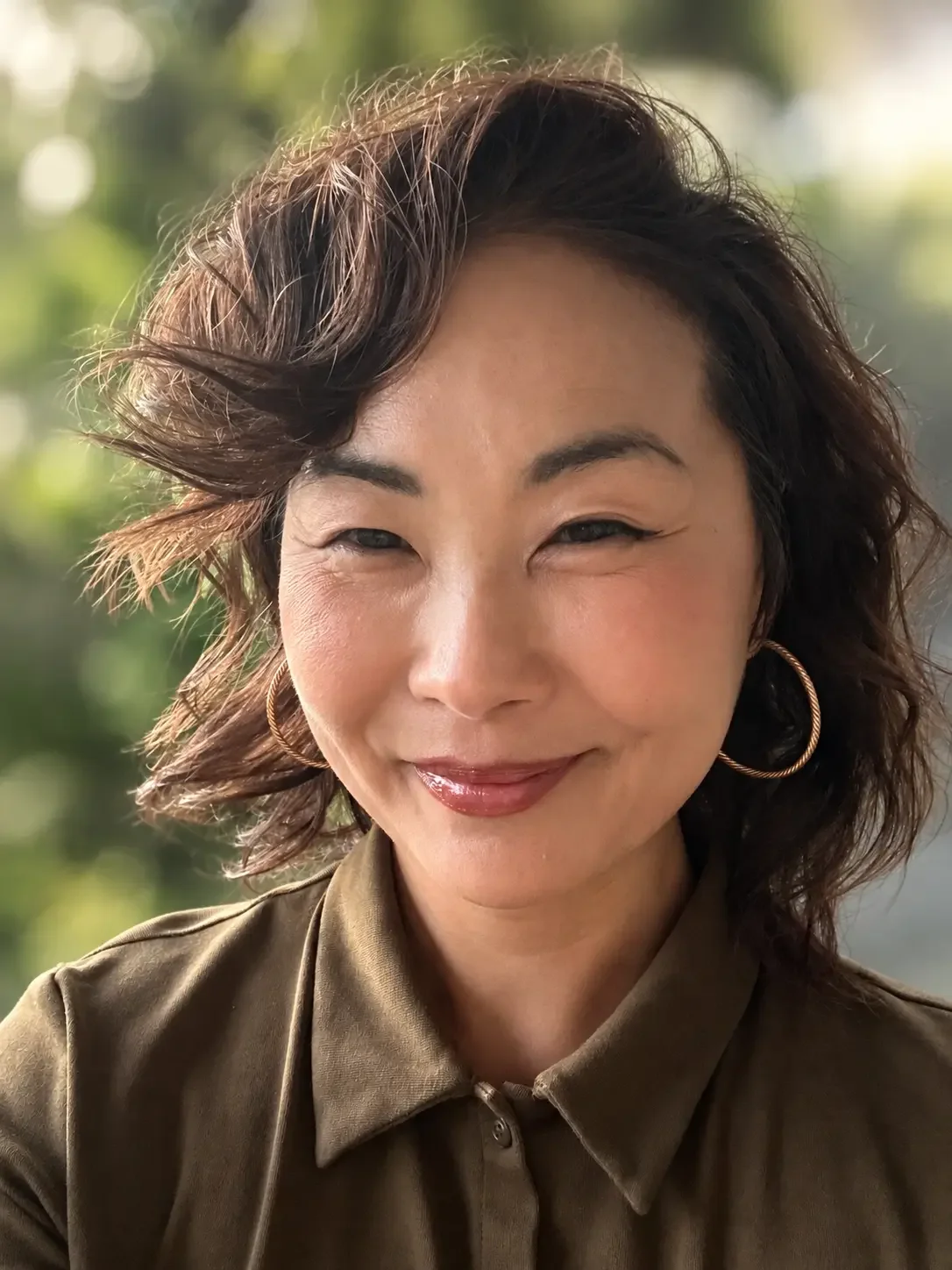 Close-up of a woman with short, wavy hair, wearing hoop earrings and a button-up shirt, smiling outdoors with a blurred green background.