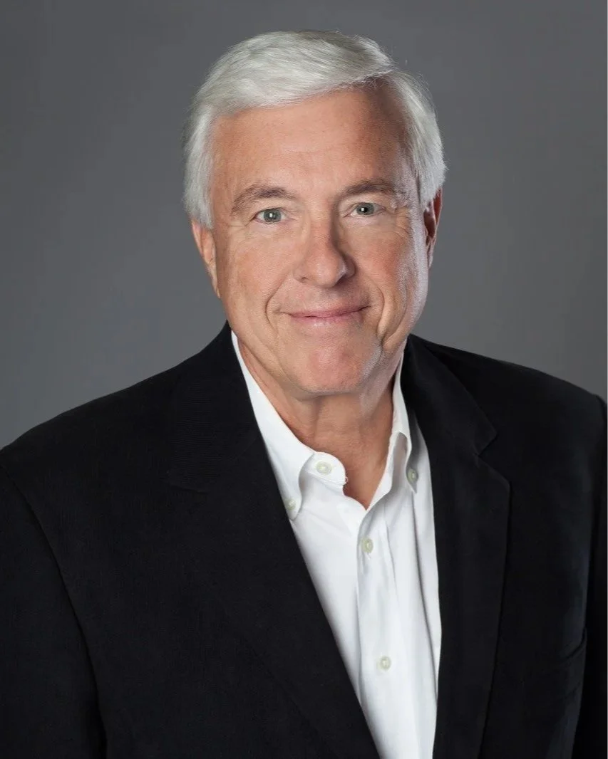 Portrait of a mature man with white hair wearing a black blazer and white dress shirt against a gray background.