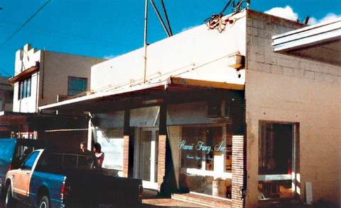 A small storefront with a glass front door and window, with a person standing outside, parked cars nearby, and a partially visible sign on the building.