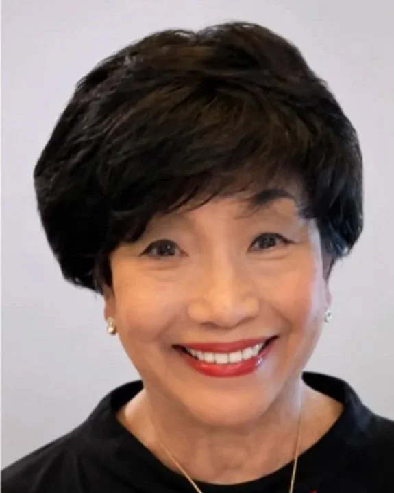 A smiling Asian woman with short dark hair, wearing pearl earrings, a black top, and a gold necklace.