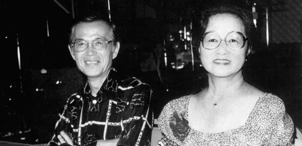 A black-and-white photo of a smiling man and woman sitting together, both wearing glasses, in an indoor setting.