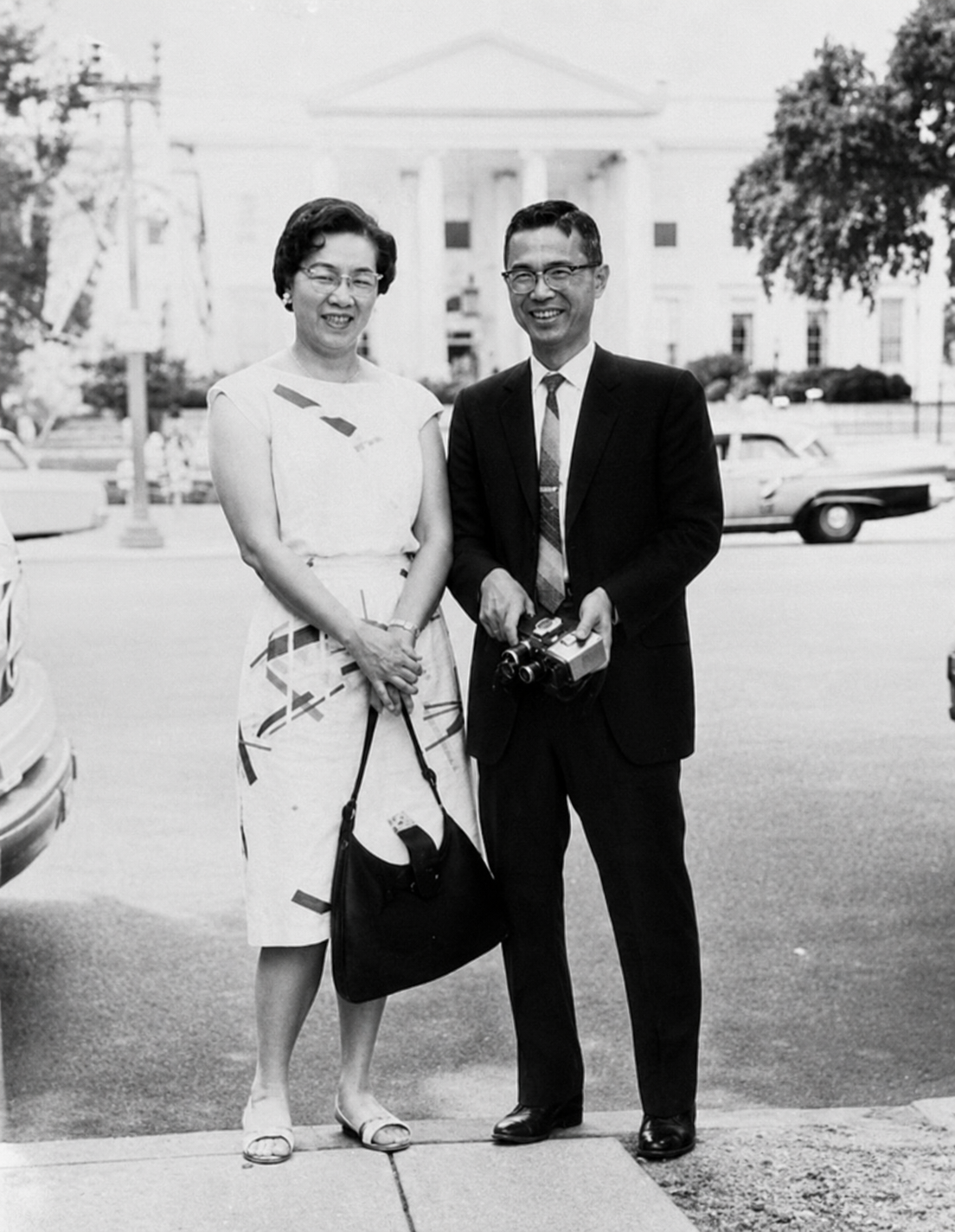 Black and white photograph of a woman and a man standing outside on a city street, smiling. The woman wears a patterned dress and carries a purse, the man wears a suit and holds a camera. There are cars and a large building in the background.