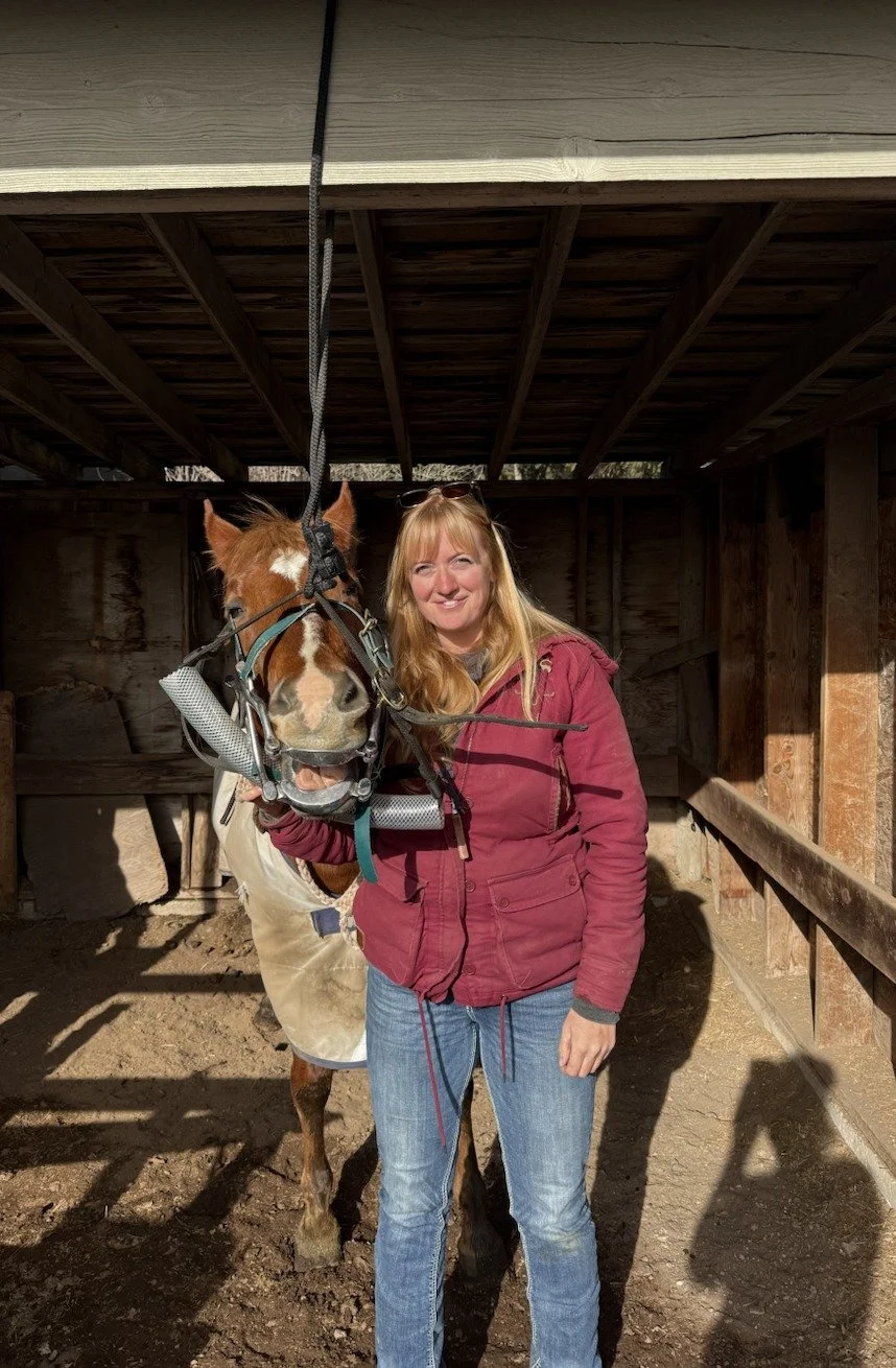 A woman with blonde hair wearing a red jacket and jeans standing next to a brown and white horse inside a barn.