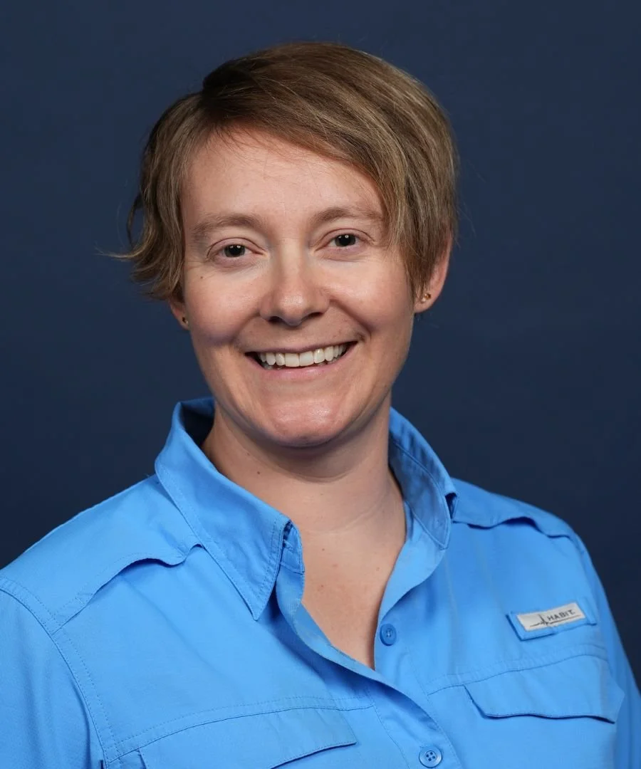 A woman with short light brown hair smiling, wearing a blue collared shirt with a name tag, against a dark blue background.