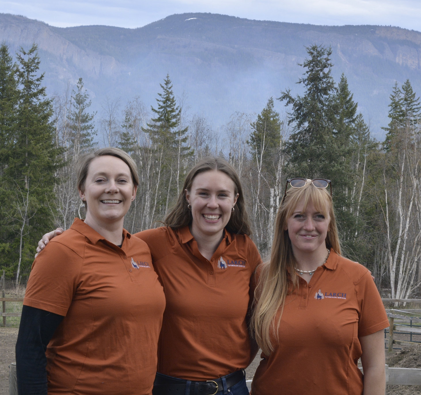 Three women in matching orange polo shirts smiling outdoors with trees and mountains in the background.